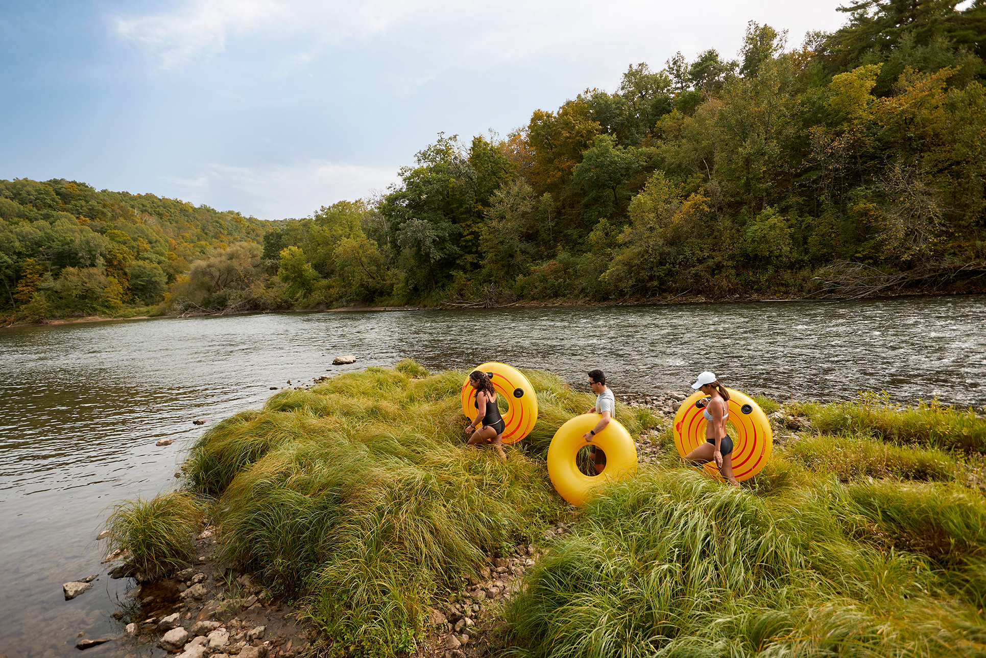 A trio of friends trek over high grass to find the perfect launch point for their bright yellow inner tubes under a light blue sky