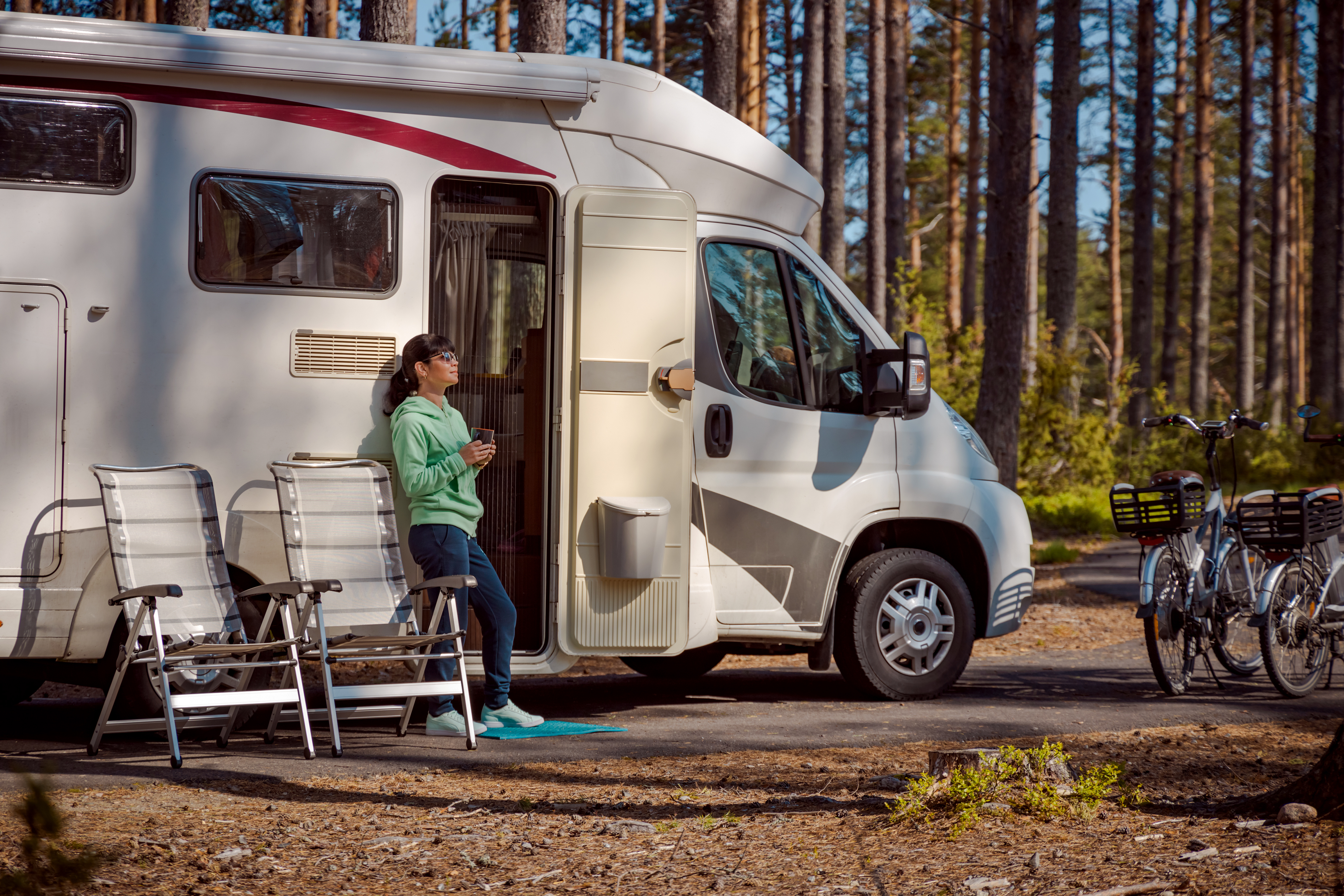 Woman stands by her RV with a cup of coffee, basking in the forest scenery