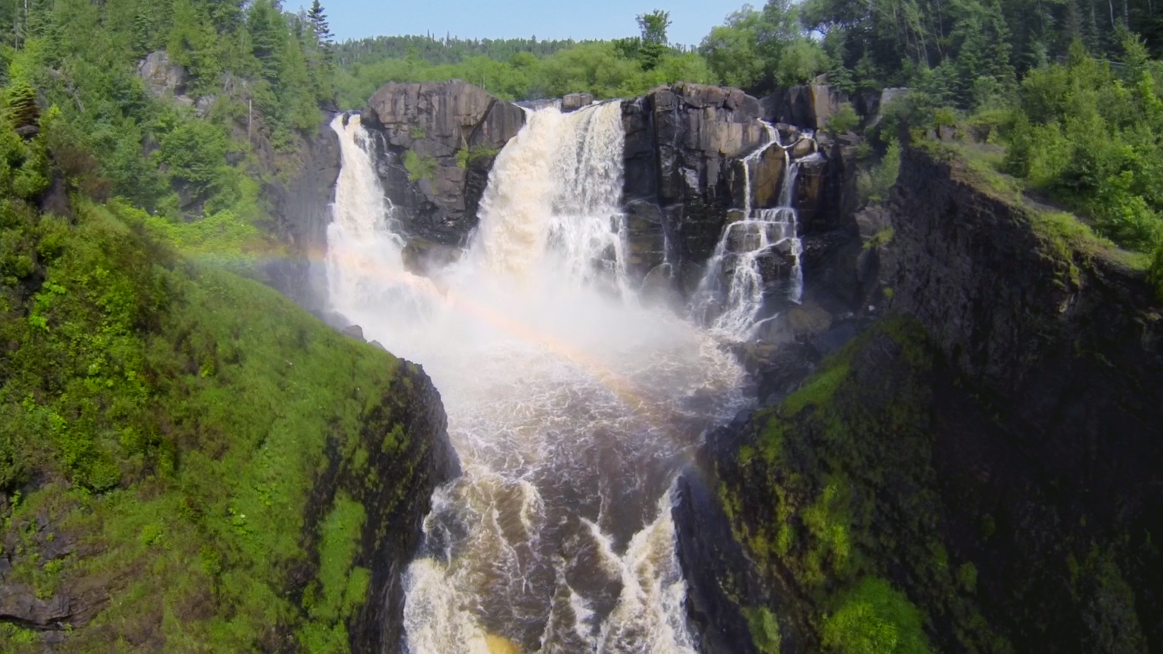Rainbow in front of a large waterfall surrounded by green trees and blue sky.