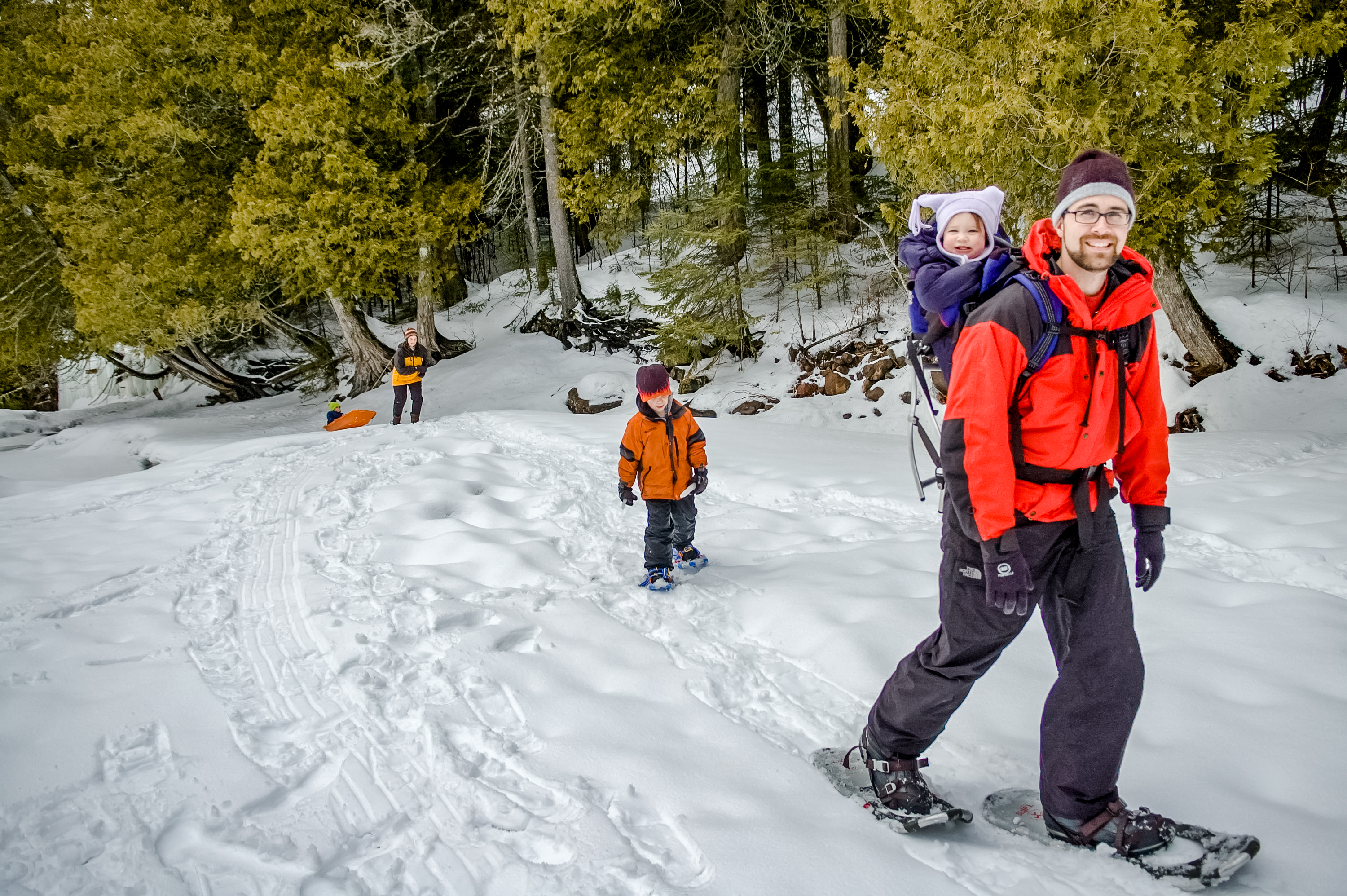 man snowshoeing with baby in backpack and young child snowshoeing behind him.