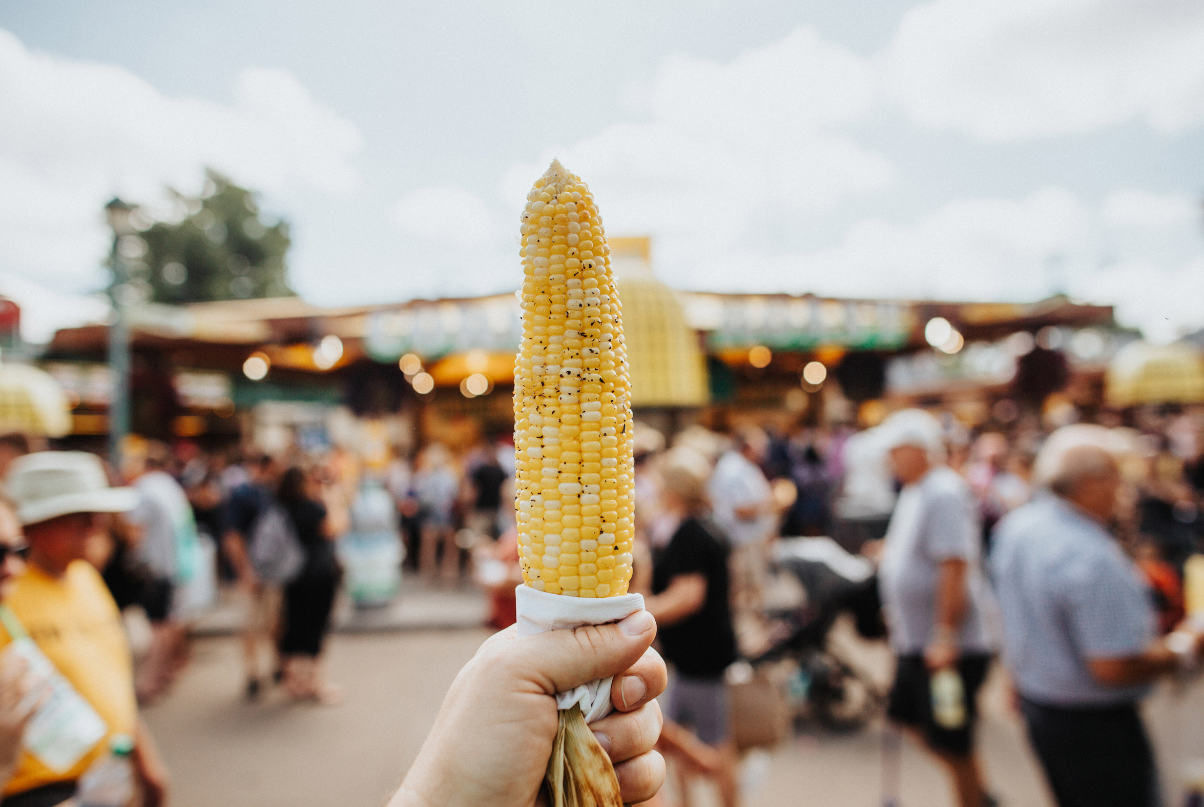 Someone holds up an ear of grilled corn in front of a State Fair crowd