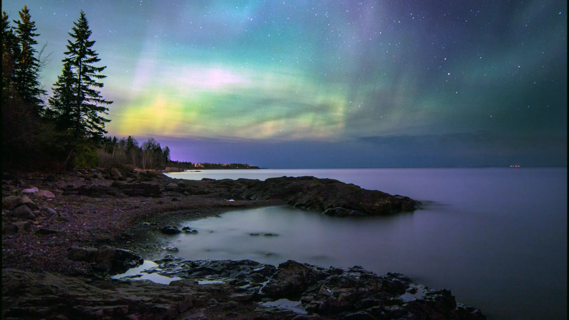 Vibrant green and purple Northern Lights streaking across a starry night sky and reflecting on the calm water of Lake Superior