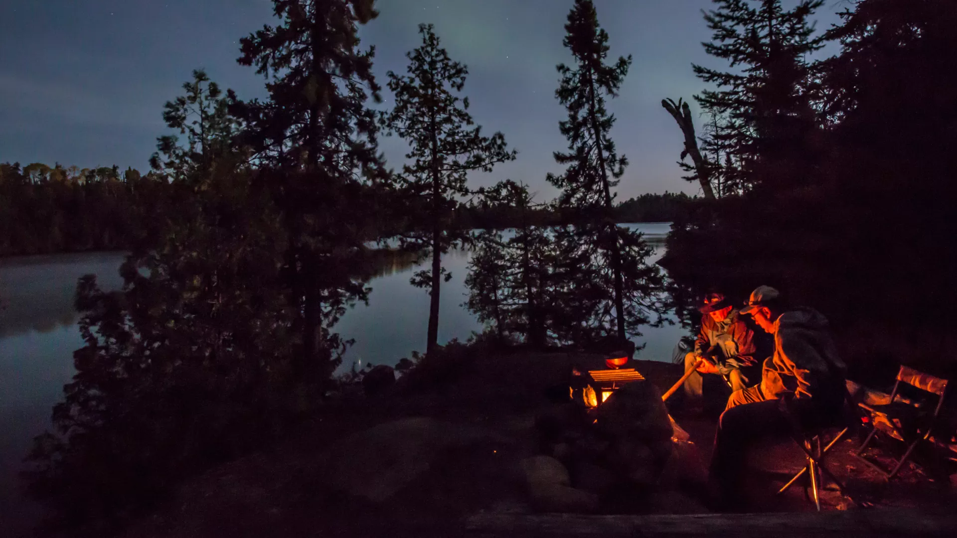 Two people sit around a campfire in the Boundary Waters as the northern lights glimmer in the distance