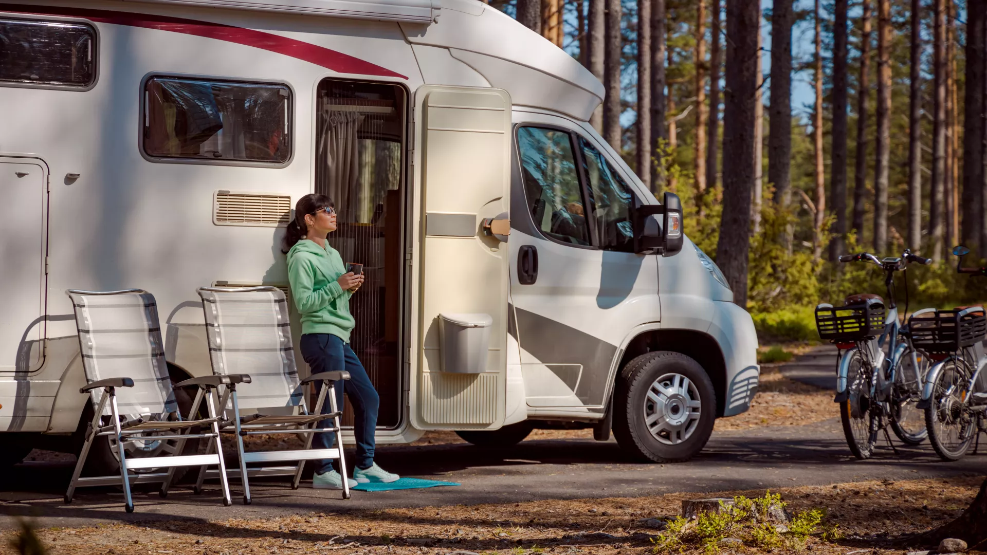 Woman stands by her RV with a cup of coffee, basking in the forest scenery