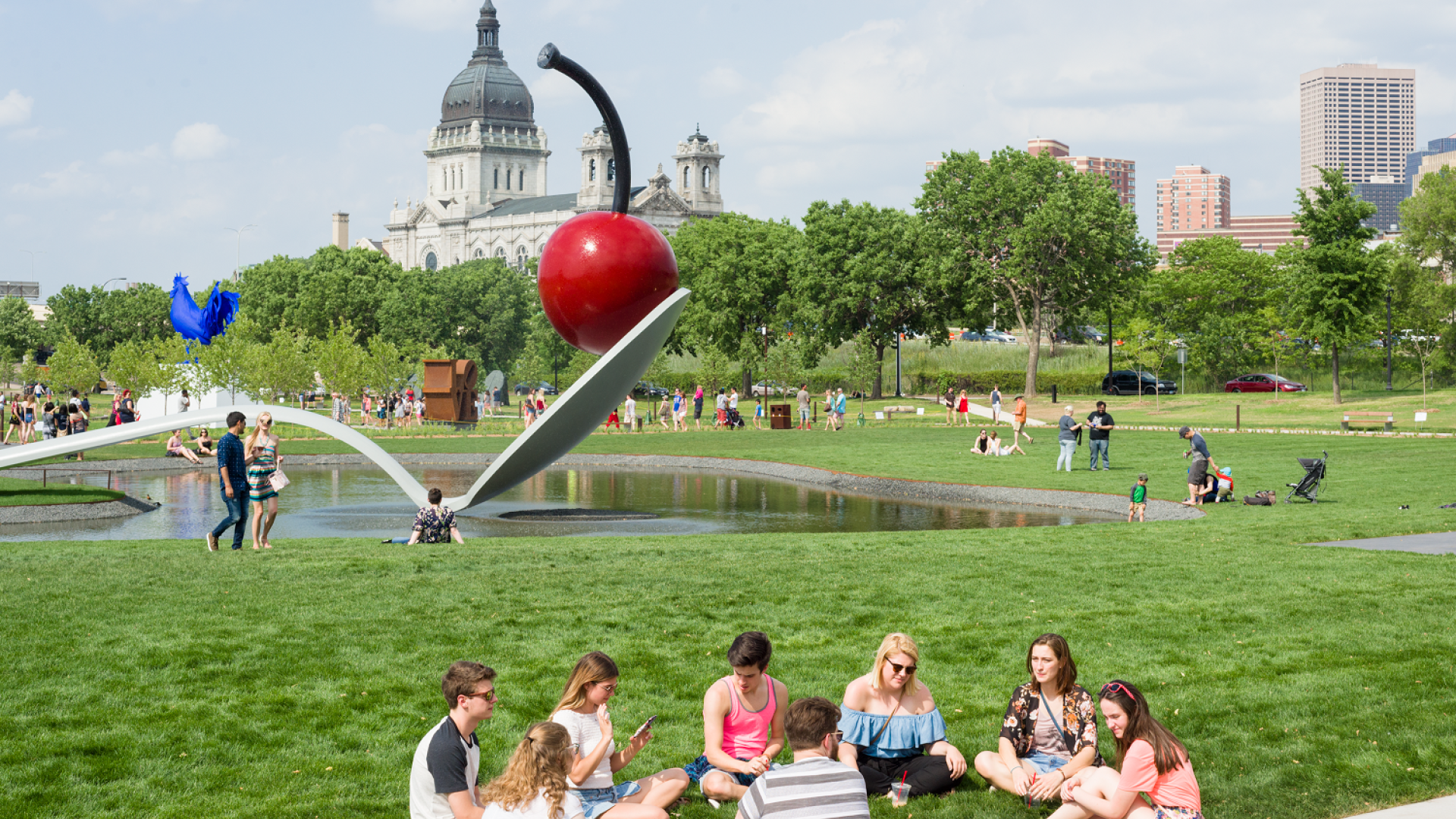 Group of people sit in a circle in front of the Spoonbridge and Cherry sculpture on a busy summer day