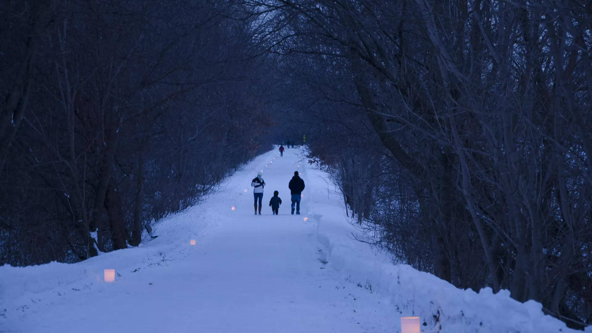 Two adults and a child walking on a snowy trail lined on both sides with luminaries