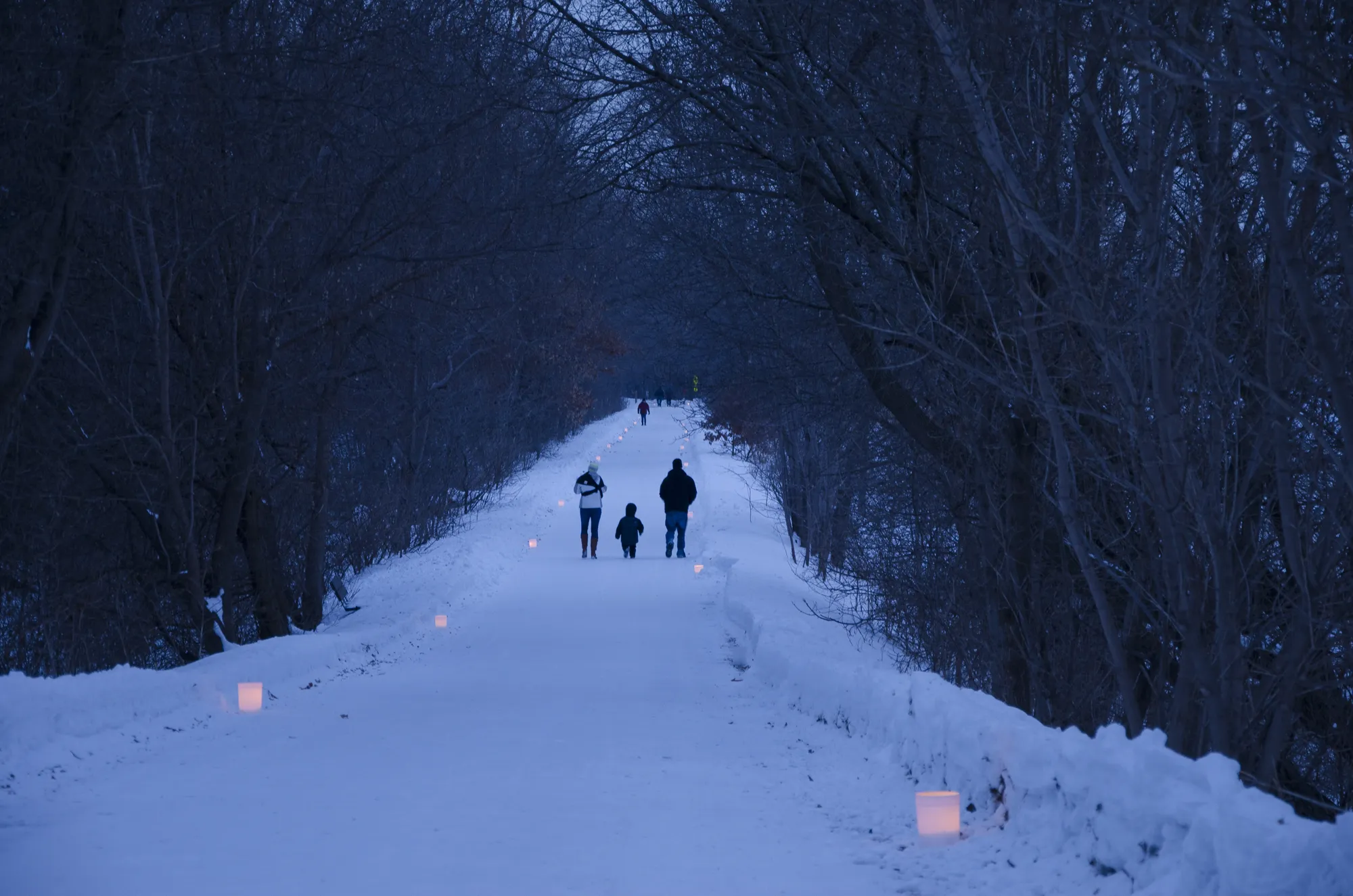 Two adults and a child walking on a snowy trail lined on both sides with luminaries