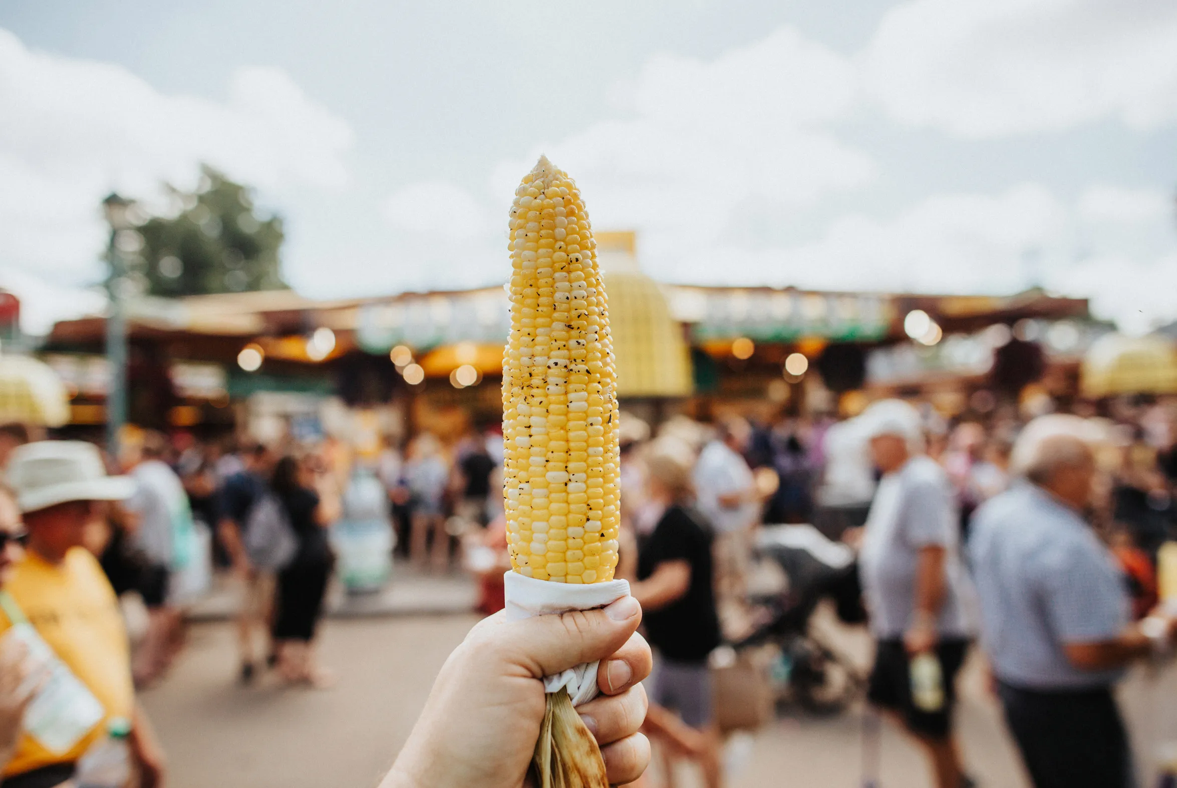 Someone holds up an ear of grilled corn in front of a State Fair crowd