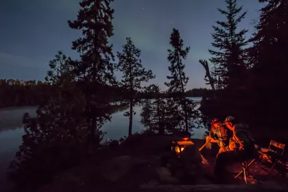 Two people sit around a campfire in the Boundary Waters as the northern lights glimmer in the distance