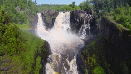 Rainbow in front of a large waterfall surrounded by green trees and blue sky.