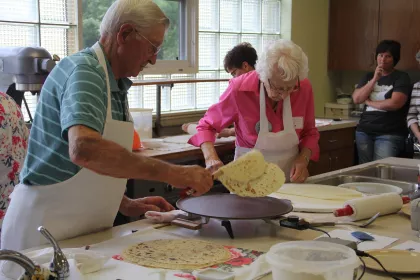 People making lefse on a cast iron griddle in a kitchen.
