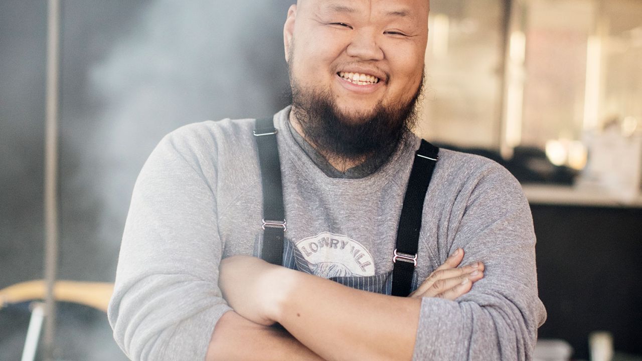 Yia Vang stands smiling, arms crossed, under a large outdoor tent with smoke from a cooking fire