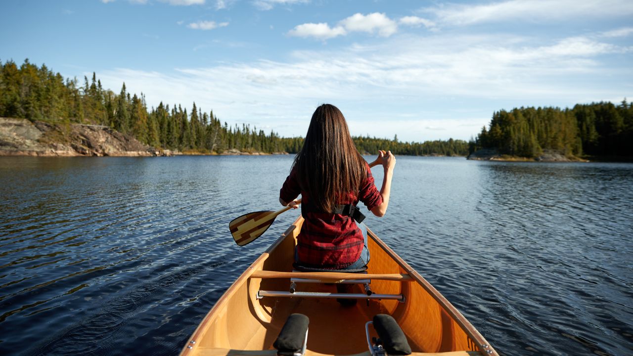 Woman in red shirt canoeing in the Boundary Waters, surrounded by dense forests and clear blue skies