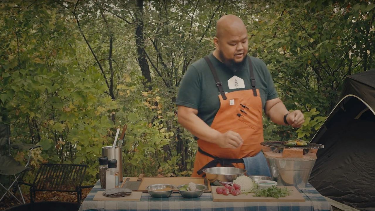 Chef Yia Vang stands at a table in a campground facing a clay pot grill with roasting jalapeños on its surface