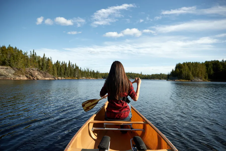 Woman in red shirt canoeing in the Boundary Waters, surrounded by dense forests and clear blue skies