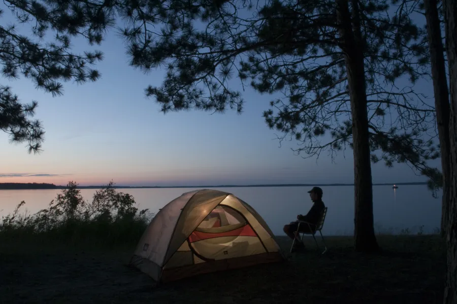A man sits by a tent near the water at night