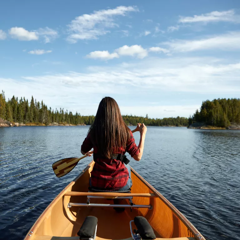 Woman in red shirt canoeing in the Boundary Waters, surrounded by dense forests and clear blue skies