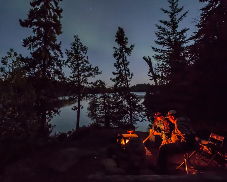 Two people sit around a campfire in the Boundary Waters as the northern lights glimmer in the distance
