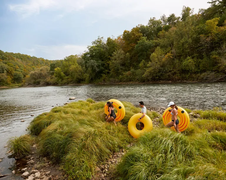 A trio of friends trek over high grass to find the perfect launch point for their bright yellow inner tubes under a light blue sky